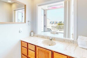 A bathroom with a sink, mirror, and window with a view of a white fence and buildings. - Bayshore 603A (Ocean City)