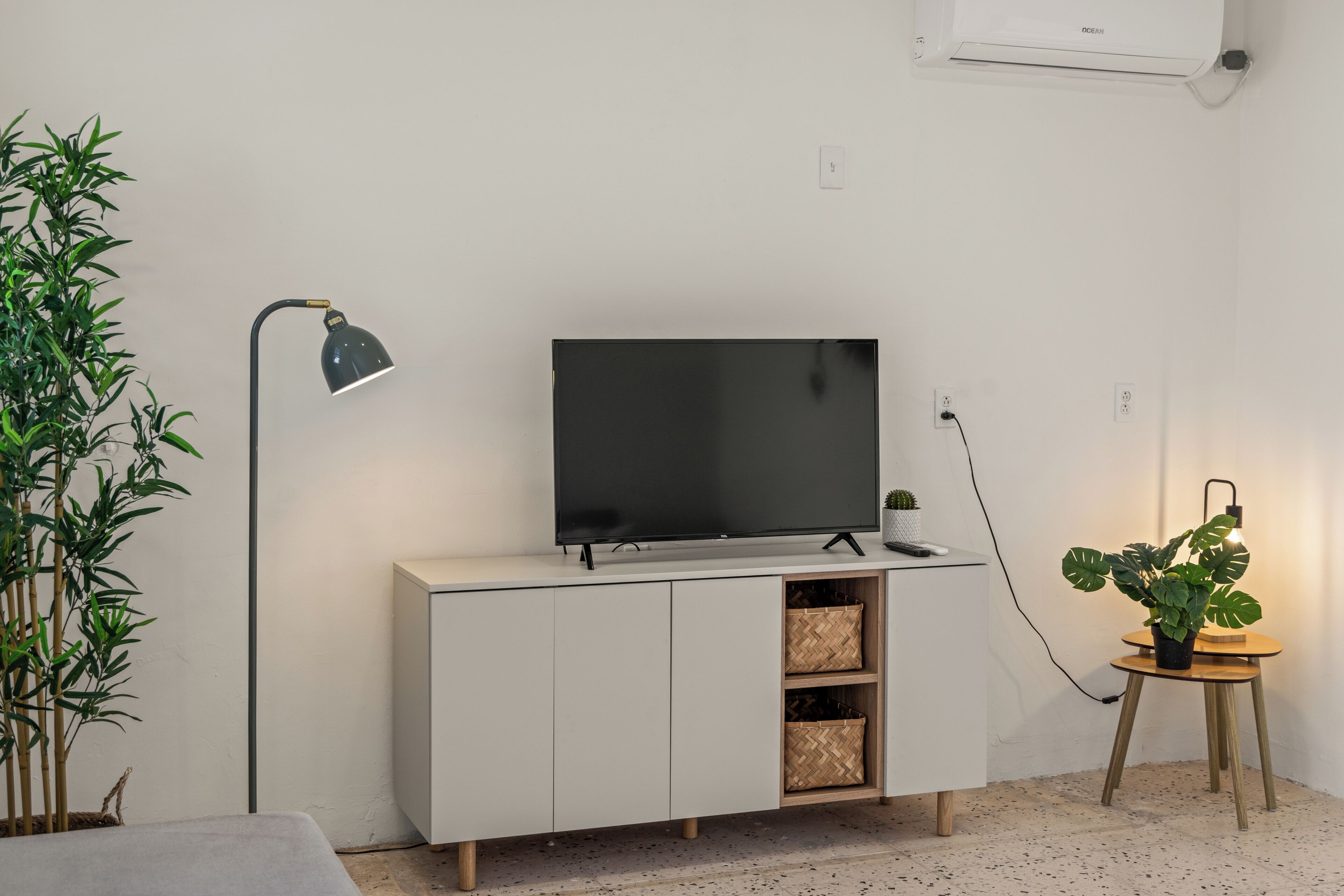 Una sala de estar moderna con una televisión de pantalla plana sobre un mueble blanco, una lámpara de pie, una planta en maceta y una pequeña mesa de madera.