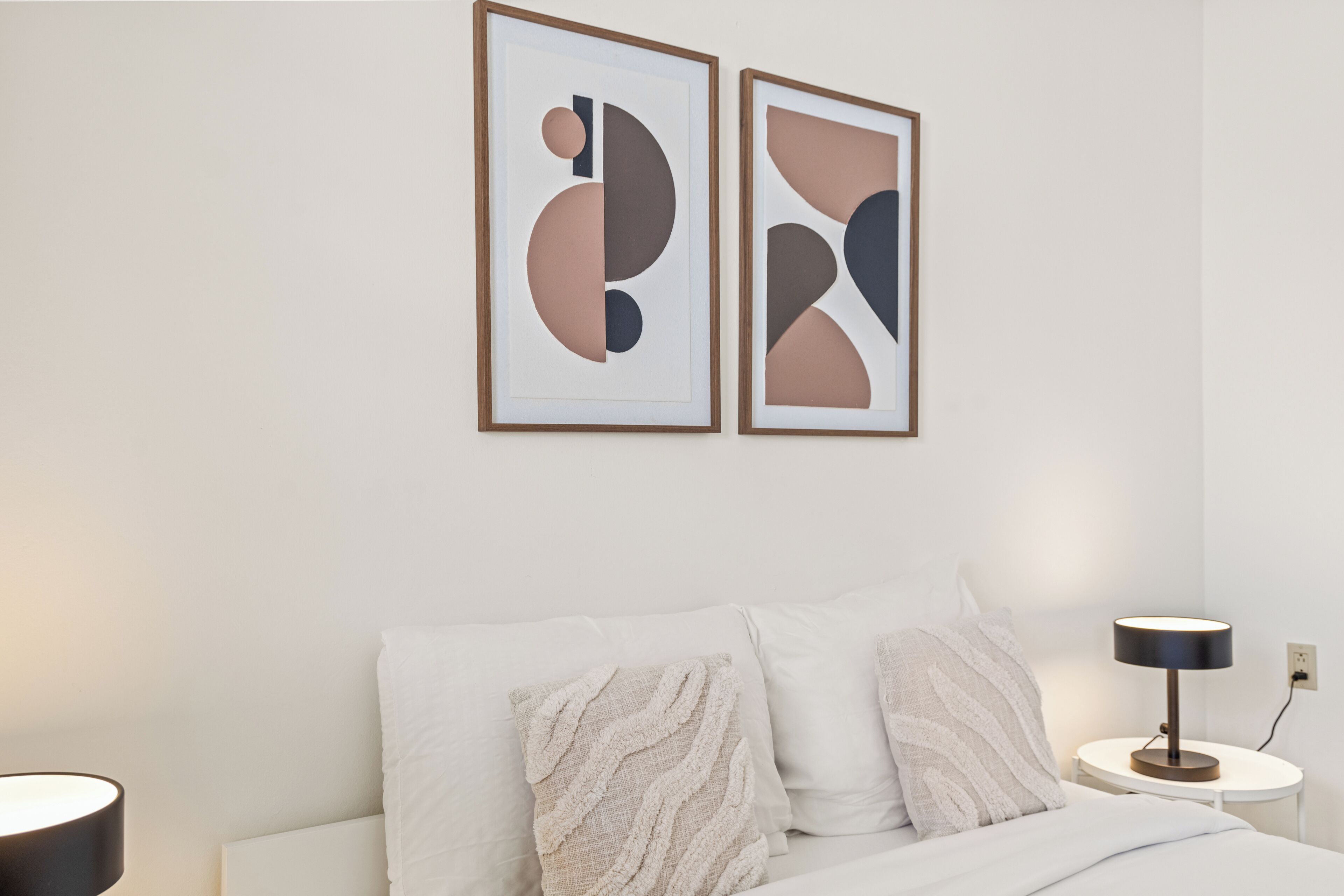 A bedroom with a bed, two pillows, a bedside table with a lamp, and two abstract paintings on the wall.