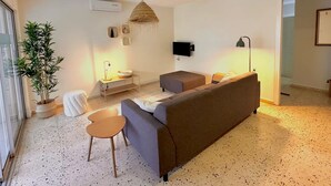 Uma sala de estar com sofá, mesa pequena, luminária, televisão e planta em vaso.