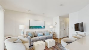 A modern living room with a sofa, ottomans, a TV, and a painting of the ocean. - Bahama Princess I (Ocean City)