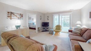 A living room with a sofa, armchair, TV, and a glass door leading to a balcony. - Coastal condo with a private washer/dryer, central AC, and a shared pool (Ocean City)