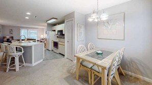 A modern kitchen with white cabinetry, a dining area with a glass table and wicker chairs, and a hallway leading to a living room. - Coastal condo with a private washer/dryer, central AC, and a shared pool (Ocean City)
