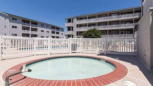 A swimming pool with a brick border, surrounded by a white fence, and adjacent to a multi-story building with balconies. - Coastal condo with a private washer/dryer, central AC, and a shared pool (Ocean City)