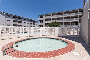 Una piscina con borde de ladrillo, rodeada por una valla blanca, junto a un edificio de varios pisos con balcones.