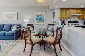 A modern kitchen with wooden cabinets, a dining area with a glass table, and a blue sofa. - Oceanfront, 13th-floor condo with pools, saunas, gym, tennis & game room (Ocean City)