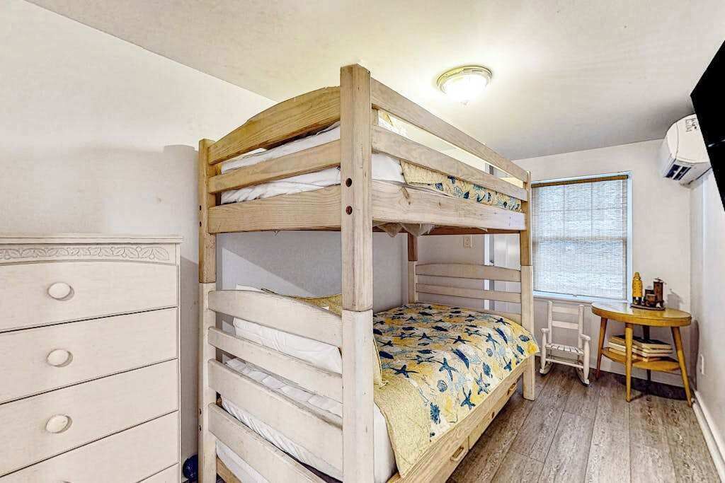 A bunk bed with a patterned bedspread, a dresser with drawers, a small table with books, and a window with blinds.