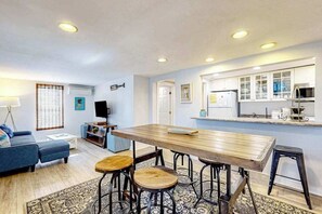A modern living room with a wooden dining table, blue sofa, and a kitchen area with white cabinets.