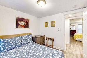 Quarto com cama, mesa de cabeceira, uma pequena mesa e uma porta que dá para outro cômodo.