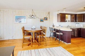 Un comedor con una mesa redonda de madera y sillas, una cocina con armarios oscuros y un microondas blanco, y una lámpara colgante.