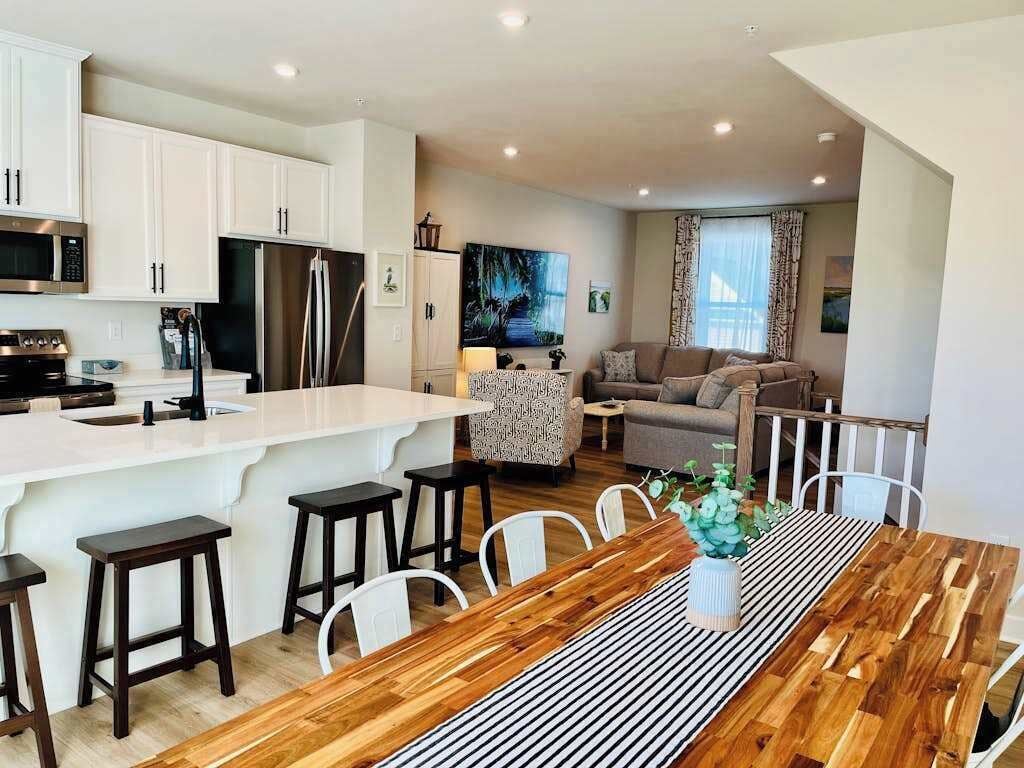 A modern kitchen with white cabinets, a stainless steel refrigerator, and a dining area with a wooden table and bar stools.