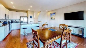 A modern kitchen with white cabinets, a dining table with wooden chairs, a TV mounted on the wall, and a kitchen island with stools. - Seaside Village Bowline 13007 -2 (West Ocean City)