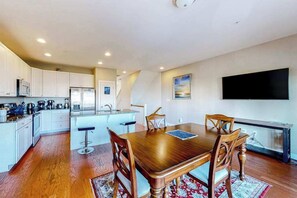 A modern kitchen with white cabinets, a dining table with wooden chairs, a TV mounted on the wall, and a kitchen island with stools. - Seaside Village Bowline  13007 -2 (West Ocean City)