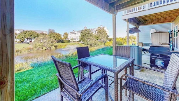 A covered patio with wicker furniture, a glass table, and a view of a pond and houses in the background. - Seaside Village Bowline 13007 -2 (West Ocean City)