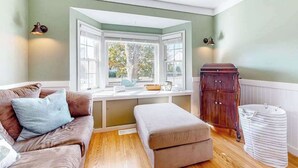 Ein Zimmer mit Erkerfenster, einem braunen Ledersofa, einem beigefarbenen Hocker, einem Holzschrank und einem weißen Wäschekorb.