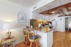 Une cuisine avec des meubles blancs, un îlot central, un coin repas avec des chaises en osier et une petite table avec une lampe.