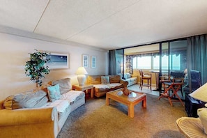 A hotel room with a sofa set, a coffee table, a dining table with chairs, a large window with curtains, and a potted plant.