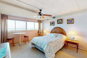 A bedroom with a bed, a ceiling fan, a window with curtains, a small table, two stools, and a nightstand with a lamp.