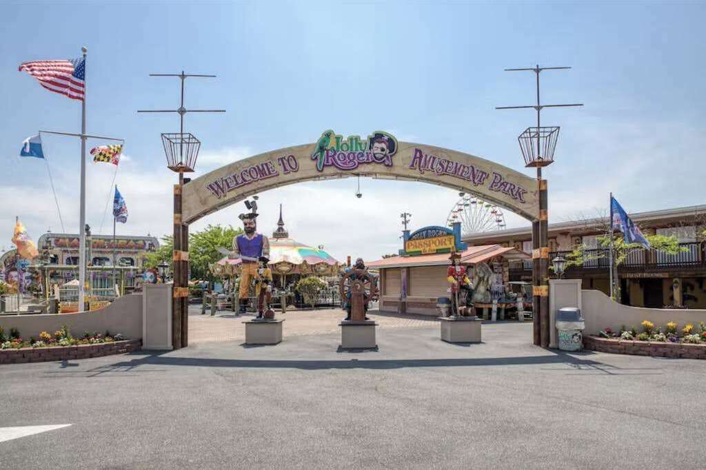 A welcome archway with 'Welcome to Rogers' and 'Rogers Amusement Park' signs, a Ferris wheel, and various flags.