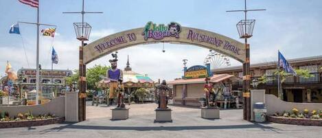 A welcome archway with 'Welcome to Rogers' and 'Rogers Amusement Park' signs, a Ferris wheel, and various flags.