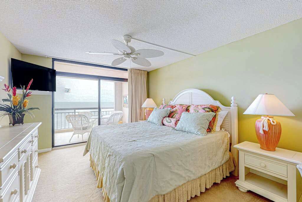 A bedroom with a bed, a ceiling fan, a nightstand, a lamp, a dresser, a TV, and a balcony with a chair.