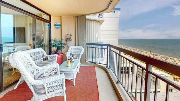 En balkon med fletningsmøbler, en rød måtte og udsigt til stranden.