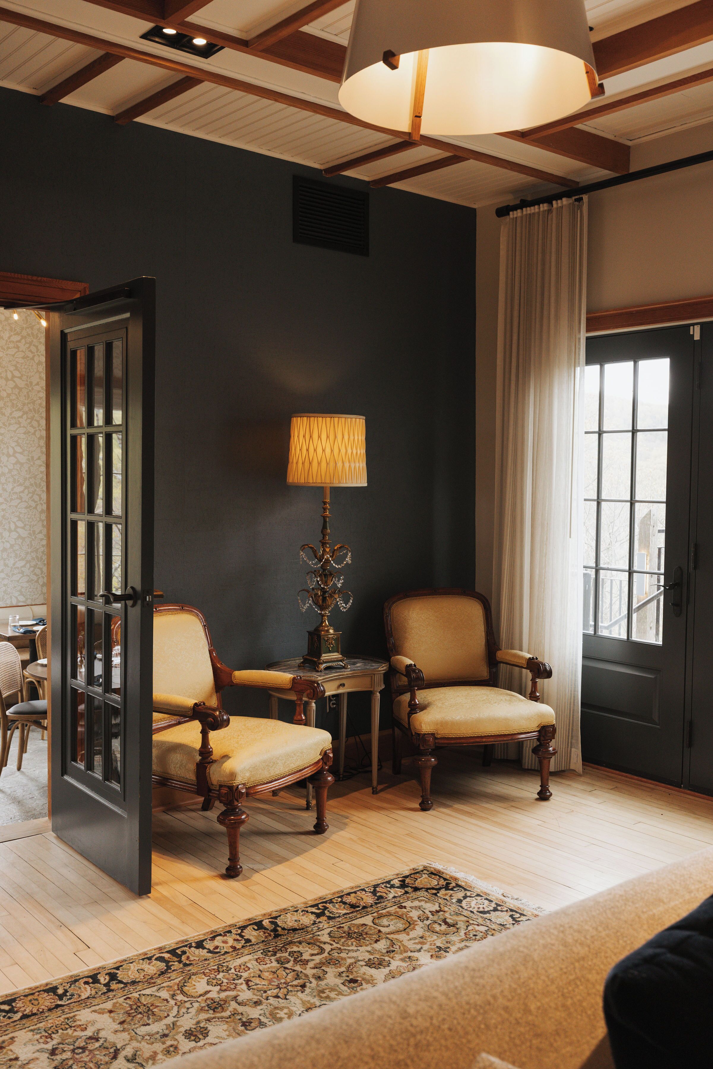 Una habitación con pared oscura, una silla de madera, una mesa con lámpara, una ventana con cortinas y una puerta.