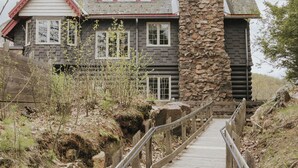 A wooden pathway leading to a log cabin with a stone chimney, surrounded by natural terrain.