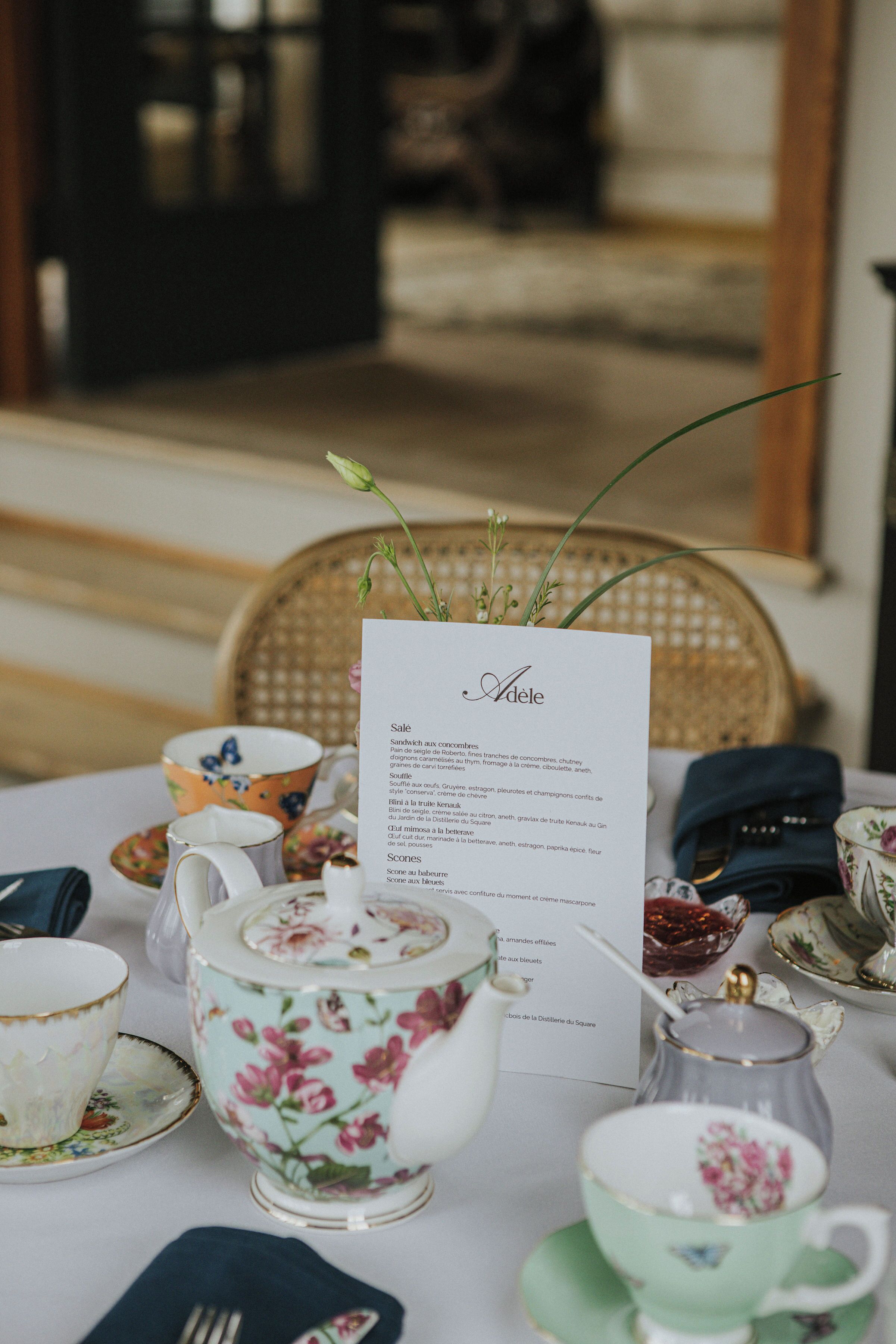Una mesa dispuesta con tazas de té de estampado floral, una tetera y un menú titulado 'Adele'.