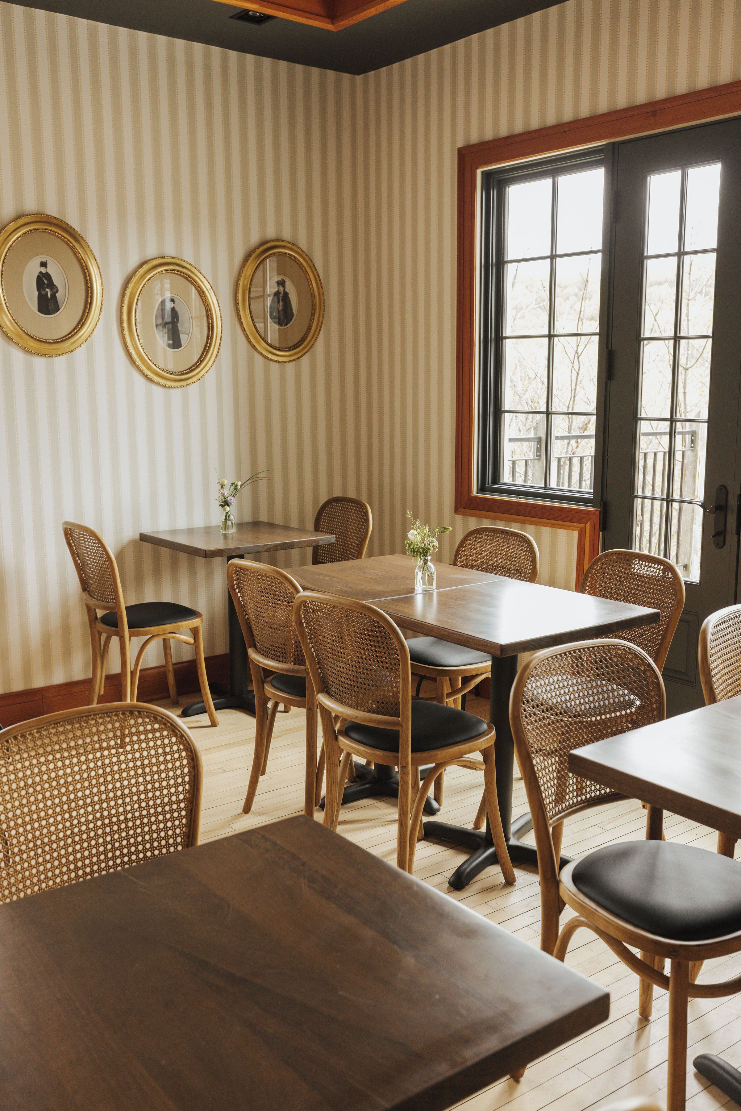 A dining area with striped wallpaper, wooden tables and chairs, and three framed pictures on the wall.