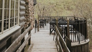 A wooden deck leading to a stone building with large windows and a view of a forested hill.