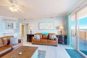 A living room with a brown leather sofa, a coffee table, a painting on the wall, and a view of the ocean through large windows. - Ocean's Edge 303 (Ocean City)