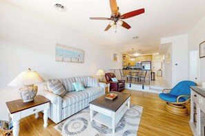A living room with a sofa, coffee table, and chairs, a ceiling fan, and a kitchen in the background. - Spacious Oceanfront - condo on the Boardwalk w/balcony, WiFi, washer/dryer & AC (Ocean City)