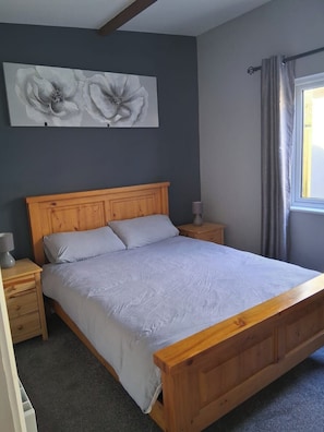 A bedroom with a wooden bed, a nightstand with a lamp, a wall-mounted artwork, and a window with curtains. - 5 bed Coastal Villa, Fethard-on-sea Hook Penisula (Saltmills)