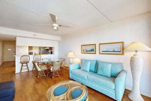 A living room with a blue sofa, a dining table with chairs, a ceiling fan, and framed pictures on the wall. - Cozy oceanfront condo with beach views, pools, gym, tennis & sauna (Ocean City)