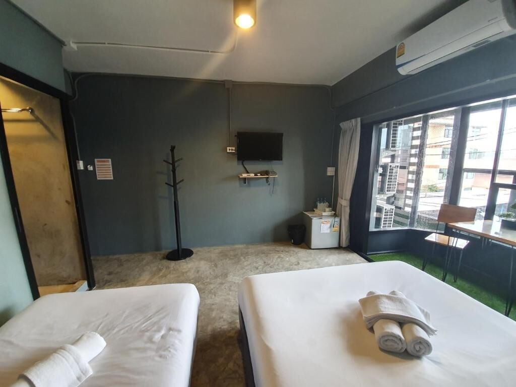 A hotel room with two beds, a TV mounted on the wall, a window with a view of buildings, and a small table with a chair.