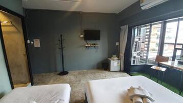 A hotel room with two beds, a TV mounted on the wall, a window with a view of buildings, and a small table with a chair.