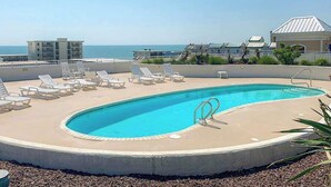 A rooftop pool with lounge chairs, a clear view of the ocean, and a building with a sloped roof in the background. - Sunny - Ocean Block Condo, steps to the beach with pool, balcony & fitness room (Ocean City)