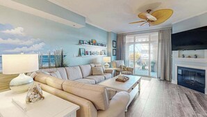 A living room with a sofa, coffee table, and a fireplace. There is a ceiling fan and a TV mounted on the wall. - Sunny - Ocean Block Condo, steps to the beach with pool, balcony & fitness room (Ocean City)