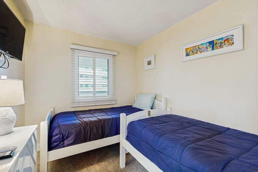 A bedroom with two beds, a window with blinds, a desk with a lamp, and a framed picture on the wall.
