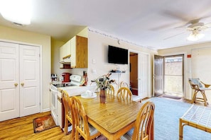 A dining area with a wooden table and chairs, a kitchen with white cabinets, a television mounted on the wall, and a ceiling fan. - Club Ocean Villa II 184 (Ocean City)