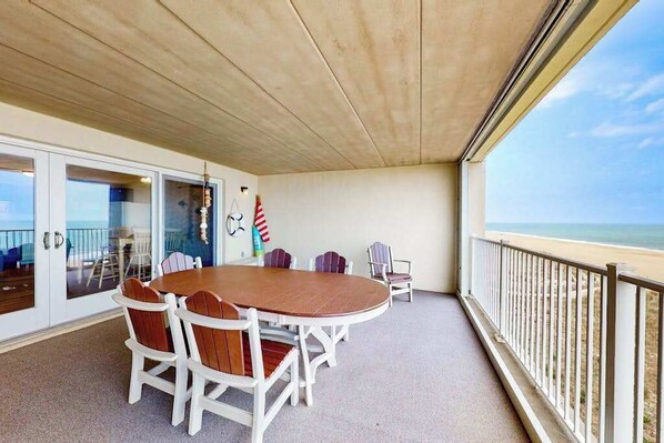 Un espacio al aire libre cubierto con una mesa redonda y sillas, vista a la playa y una barandilla.