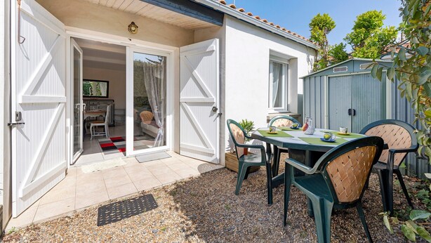 Un patio con mesa y sillas, una puerta corrediza de vidrio que da a una habitación, y un pequeño jardín.