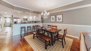 A spacious dining area with a wooden table and chairs, a chandelier, and a kitchen in the background. - 2nd-floor condo near the beach with pool, balcony, gym, fireplace, & jetted tub (Ocean City)