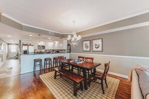 A spacious dining area with a wooden table and chairs, a chandelier, and a kitchen in the background. - 2nd-floor condo near the beach with pool, balcony, gym, fireplace, & jetted tub (Ocean City)