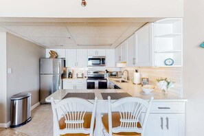 A modern kitchen with white cabinetry, stainless steel appliances, and a dining area with white chairs. - Beachfront condo with ocean views, private balcony, central AC, & W/D (Ocean City)