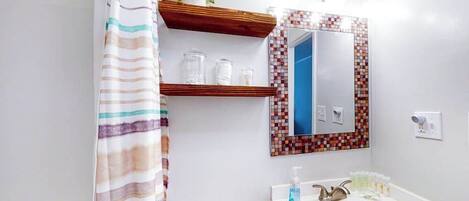 A bathroom with a striped shower curtain, a white sink, a mirror with a mosaic tile frame, and a wooden shelf with a plant.