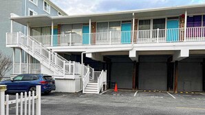 A multi-story motel with a staircase leading to the upper floor, a parking area with a blue car, and a white picket fence. - Ocean Waye 226 (Ocean City)