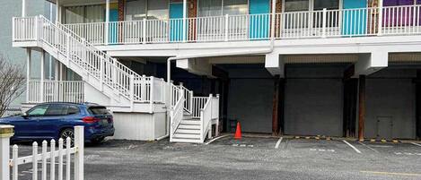 A multi-story motel with a staircase leading to the upper floor, a parking area with a blue car, and a white picket fence.