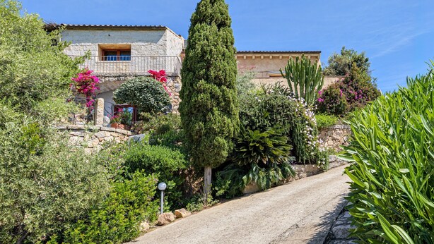 Una casa de piedra con balcón, rodeada de exuberante vegetación y un camino de grava.